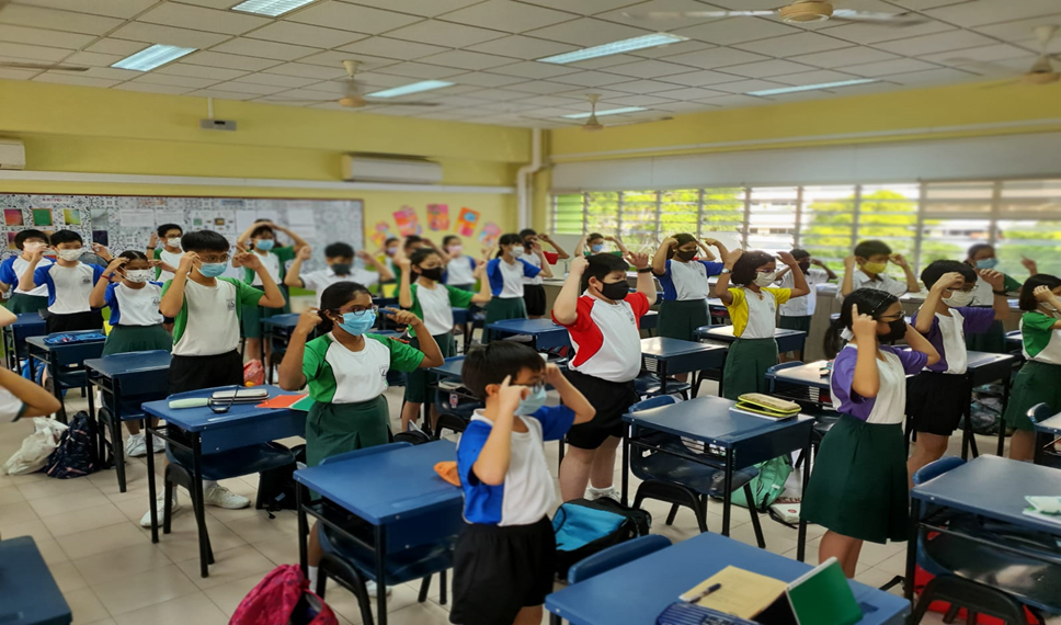 Masked students stand beside desks in a classroom, raising hands to their heads during an activity.